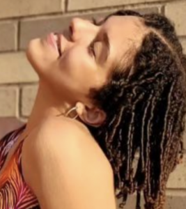 Molly Rufus has shoulder length dark finger coiled hair and a medium-light skin tone. She wears a brightly patterned halter top and hoop earrings. Her head is tilted back over her shoulder and she is shot from the left side.