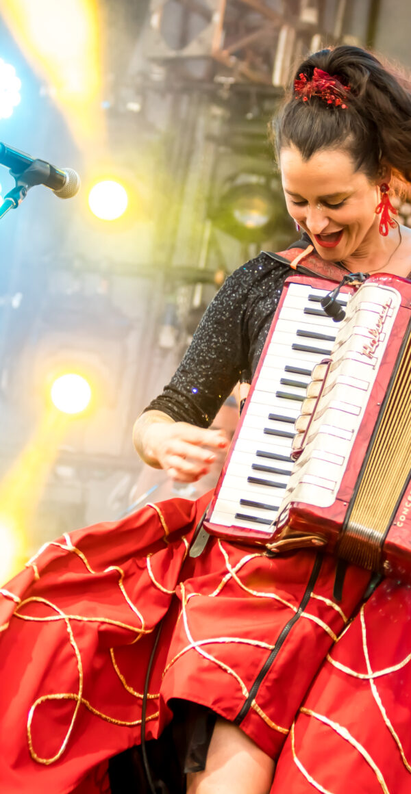 Color photo of a woman joyously playing an accordion on a large outdoor stage. To her left, a figure in a skeleton costume sits.