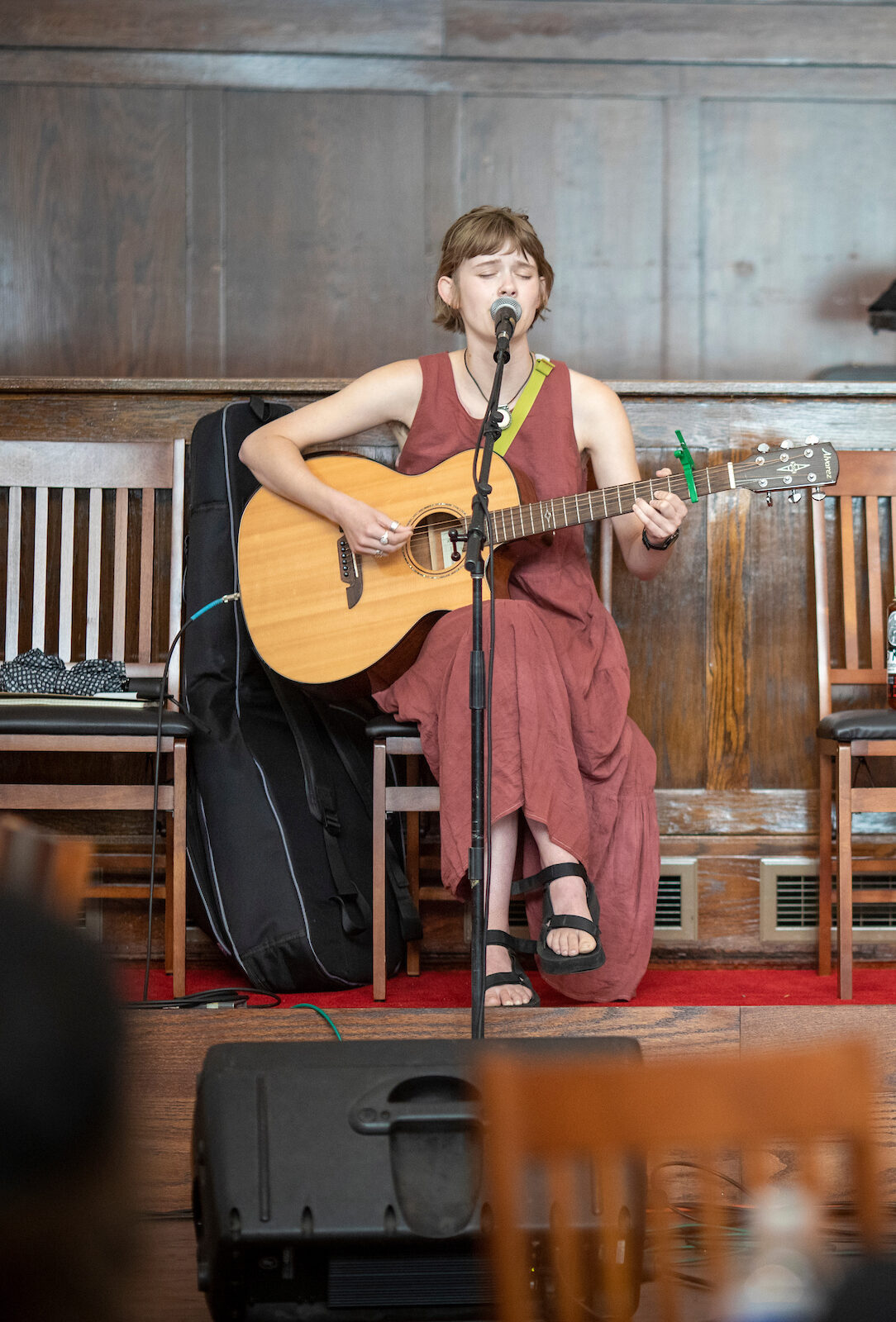 A woman sits on a wooden stage with a guitar in her arms as she sings into a stand microphone.