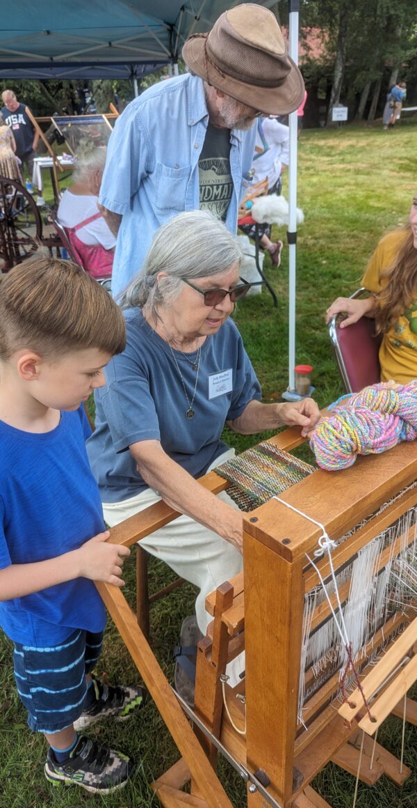 A woman sits at a loom outside and demonstrates how to weave. People stand around watching her work.
