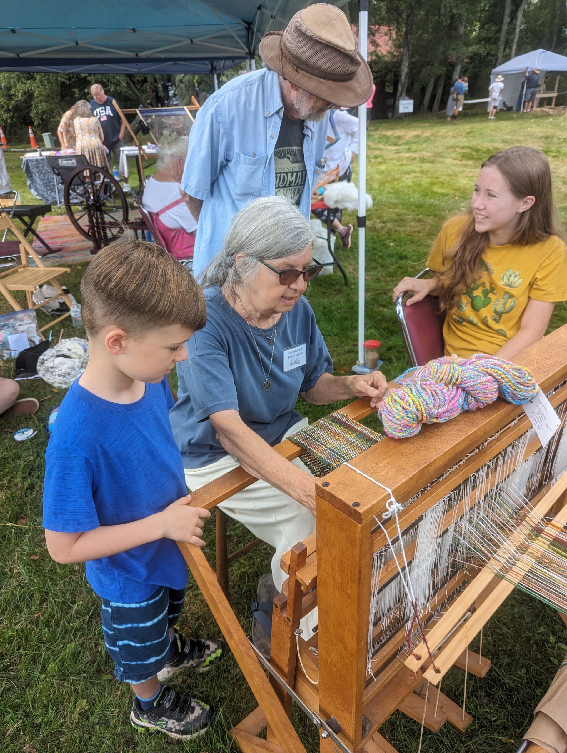 A woman sits at a loom outside and demonstrates how to weave. People stand around watching her work.