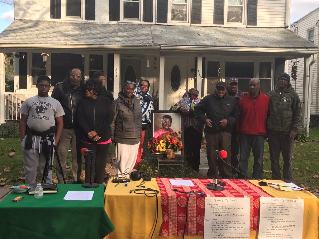 Color photo of a family outside of a large white two story clapboard house getting ready to record audio.