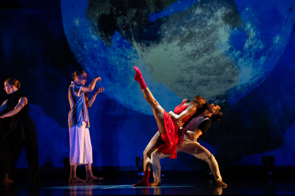 Color photo of dancers on stage with a large moon projected behind them. Center, a woman lifts her leg while a man braces her up from behind.