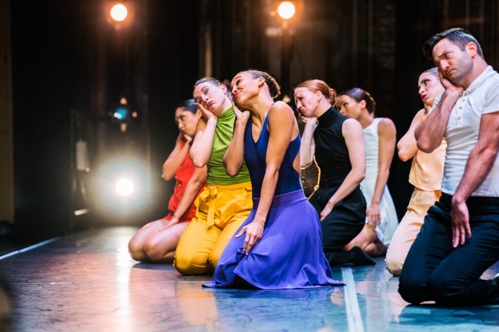 Color photo of a group of dancers kneeling onstage their heads tilted to the right into their raised hand.