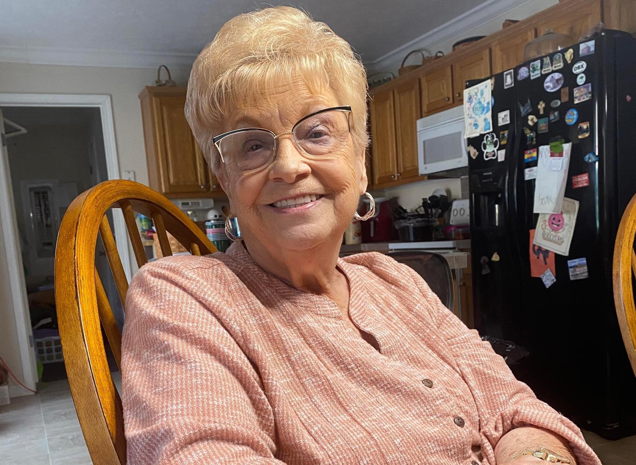 Bea Sias sits in a chair in her kitchen. She wears a pink top and has short blond hair.