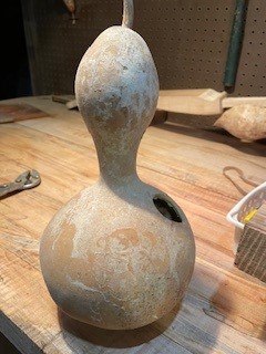 A bulbous dried gourd sits on a table. It has been scraped clean and hollowed out with a hole in the middle.