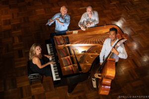 Group shot of four musicians shot from above. The musicians hold their instruments and are shot from above around a large grand piano.
