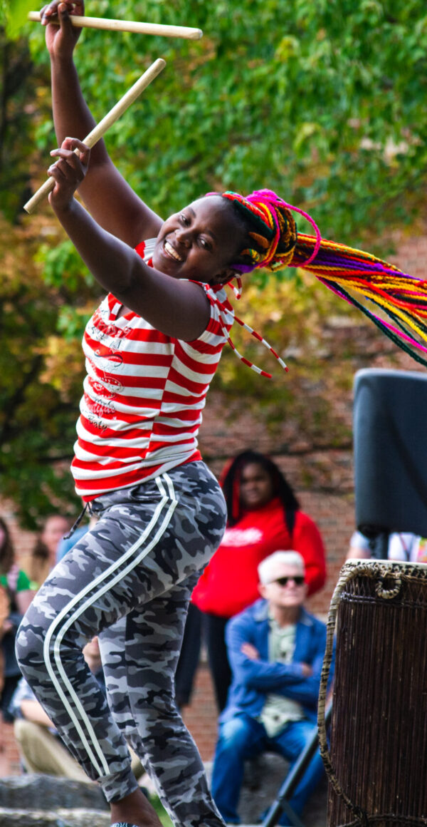 A young woman dances joyously on an outdoor stage holding drum sticks over her head.