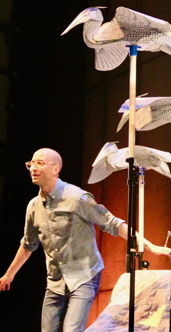 Seven large paper blue heron sculptures fill the stage space in various poses atop music stands while the performer, dressed in blue, makes a bird-in-flight pose near a blue quilt.