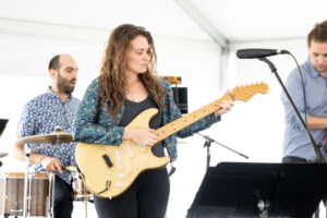 Close up of a woman playing the electric guitar with two band mates visible behind her.