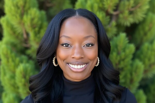 AliShah Watson has medium brown skin and long wavy hair. AliShah wears a long sleeve black turtleneck and gold and pearl earrings and is photographed in front of green foilage.