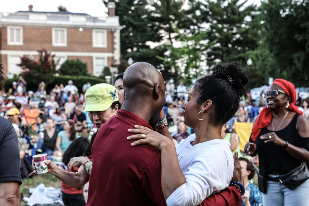 A couple dances while in the background a lawn full of audience members can be seen.