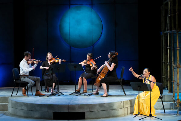 A quartet plays on a stage under a projected image of the moon. Beside them, a seated narrator tells a story.