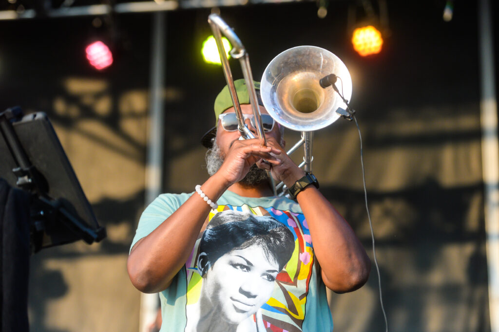 A musician plays the trombone with an outdoor stage visible behind them.
