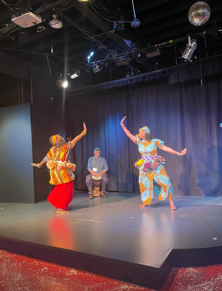 Two dancers in traditional costumes performs while a musician plays a drum in the background.