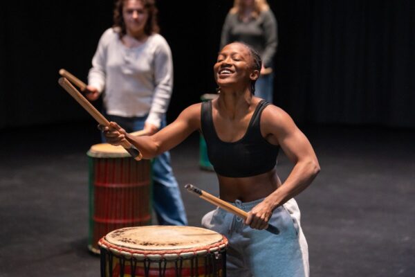 A drummer holds two drumsticks while playing a large drum. Behind them, other drummers can be seen.