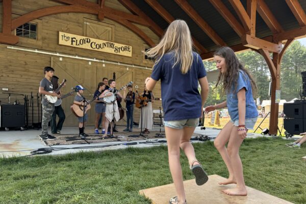 Two young people dance on a wooden board wjile a band plays in an outdoor pavilion in front of them.