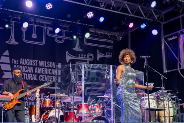 A woman stands center stage in front of a stand mic. Behind her, musicians perform on a brightly lit stage.