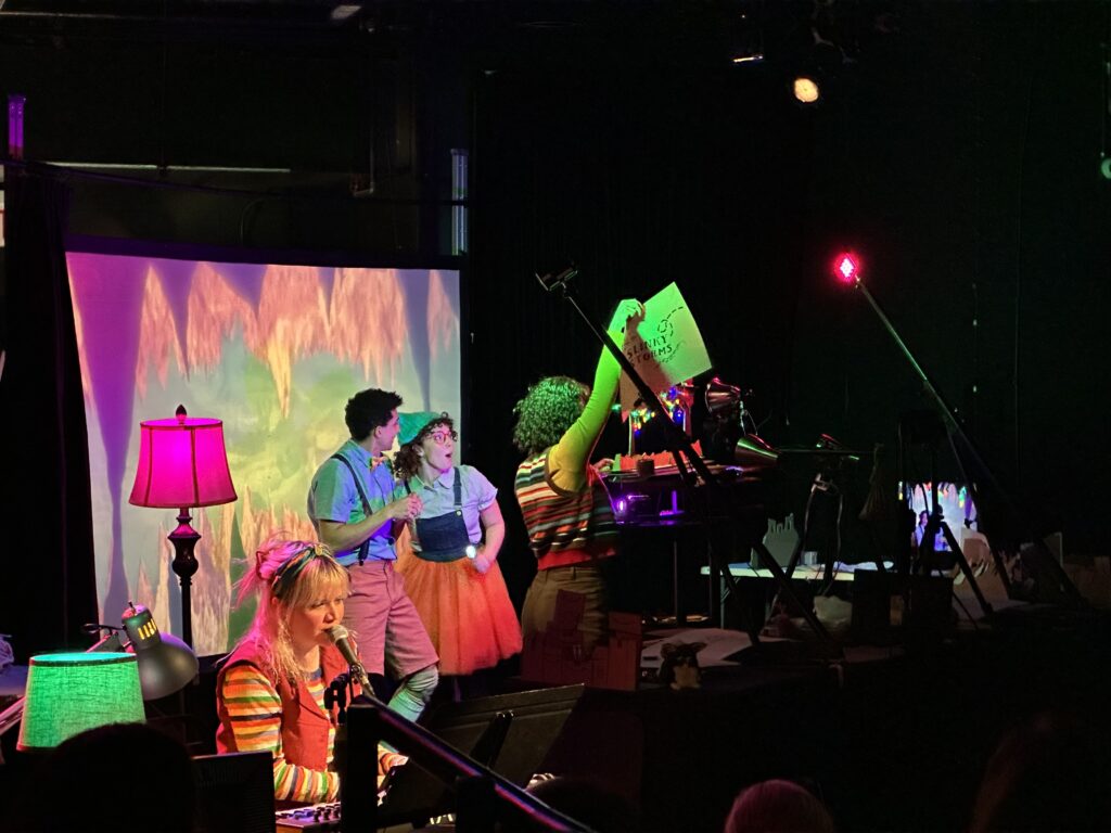 An onstage performance with two actors center and visible performers holding up cue cards and speaking into a mic.