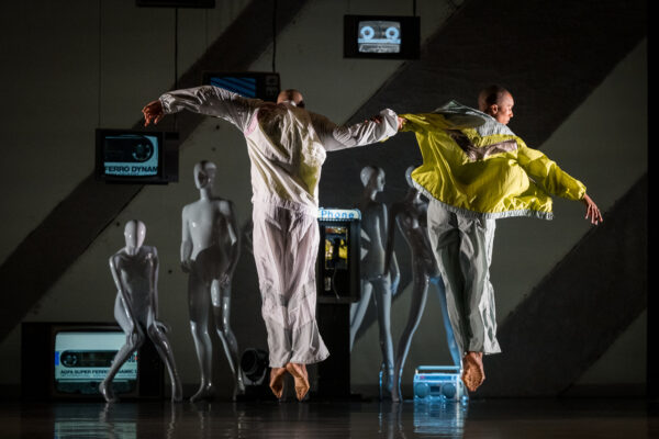 From the back, we see two dancers leap. Toes pointed down they hold arms in the center with the other arm outstretched. In the background, mannequins stand and tape cassette can be seen projected on the wall.