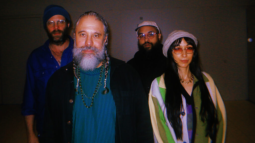 Four people slightly smiling at the camera. Three men and one woman; two men and the woman are wearing hats. The founder, Joshua Abrams, has braided pigtails and is wearing a beaded necklace.