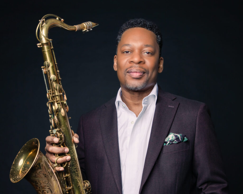 Ravi Coltrane stands facing the camera and lifts his sax in his right hand.