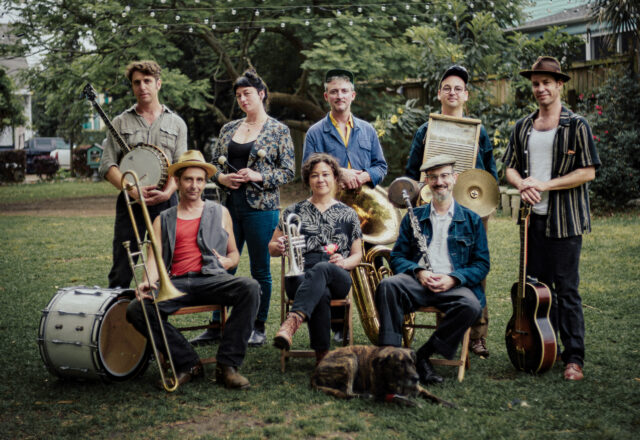 A group of eight musicians stand and sit outside holding their instruments in their hands.