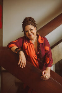 Fabiana Cozza stands on a staircase with both hands in front on the rail facing out. She wears a brightly patterned rust and orange blouse.