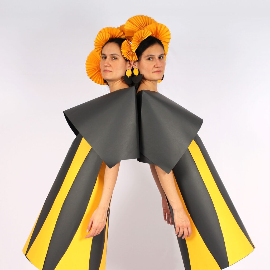 Two women stand back to back and glance sideways at the camera. They wear grey and yellow striped dresses with large grey collars and orange headdresses.
