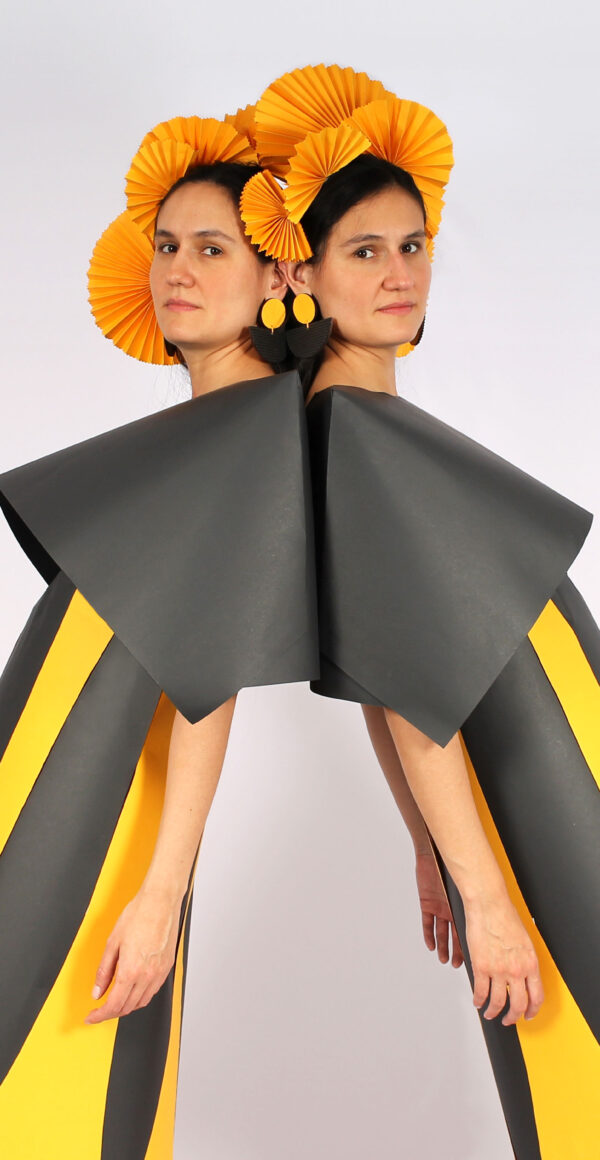Two women stand back to back and glance sideways at the camera. They wear grey and yellow striped dresses with large grey collars and orange headdresses.