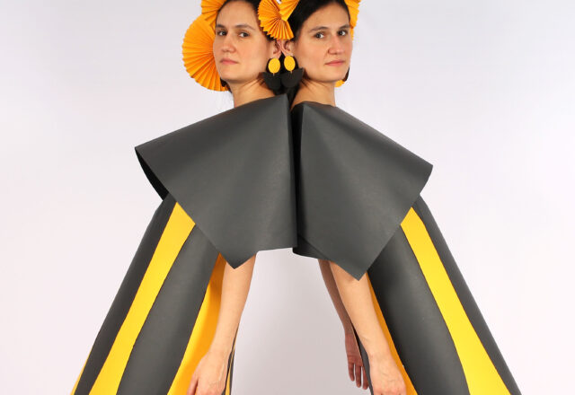 Two women stand back to back and glance sideways at the camera. They wear grey and yellow striped dresses with large grey collars and orange headdresses.