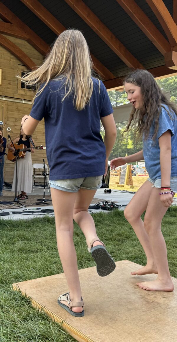 Two girls clog on a board in the grass while musicians play on a stage.
