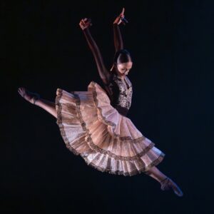 A dancer leaps through the air arms above her head and ruffled skirts flaring.