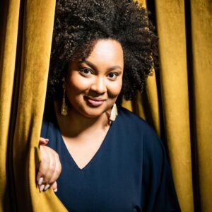 Lucibela peers out from between two gold stage curtains.