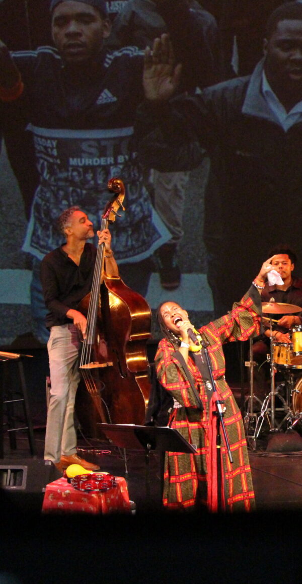A woman sings into a mic center while a bassist and percussionist play behind her.