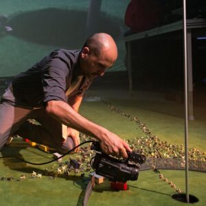 A man kneels with a video recorder on a green carpet filming small plastic figures heading into a golf course hole.