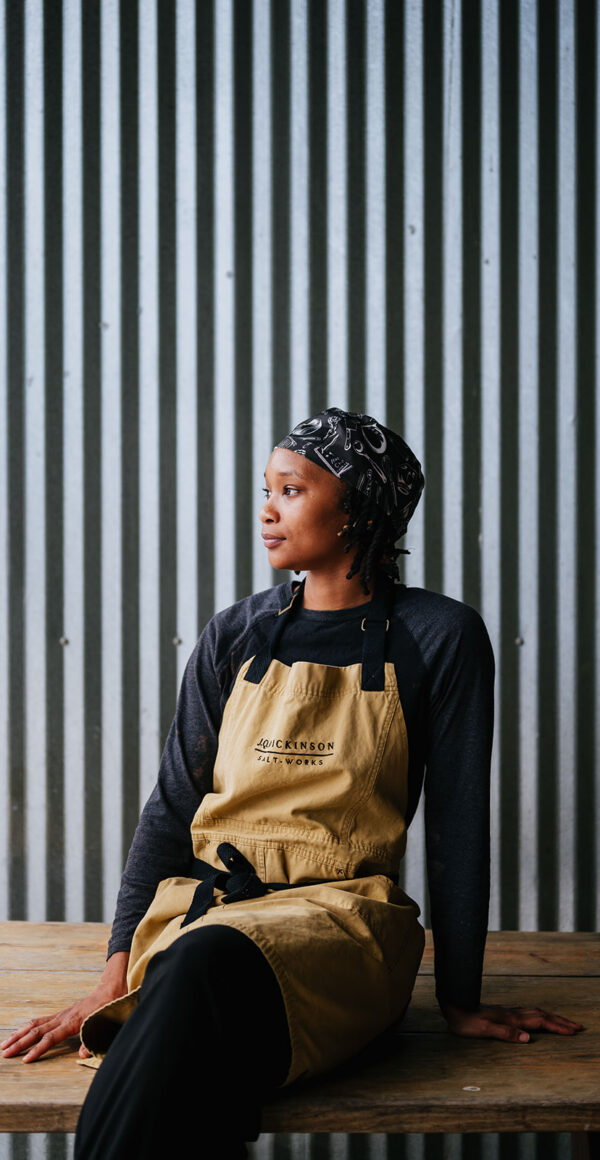 Keyarna “Chef Ke” Frederick sits on a wooden table with corrugated steel behind them. They wear a chefs apron and head scarf.