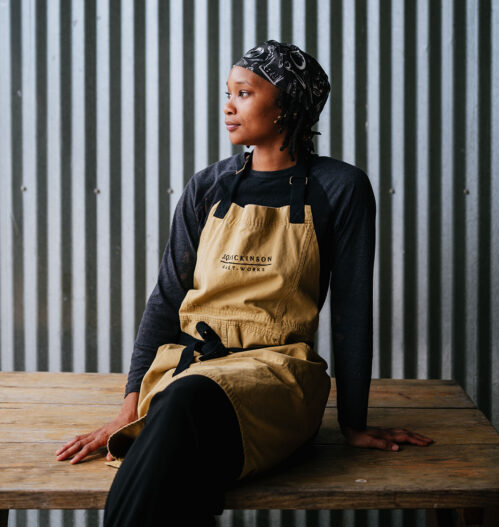 Keyarna “Chef Ke” Frederick sits on a wooden table with corrugated steel behind them. They wear a chefs apron and head scarf.