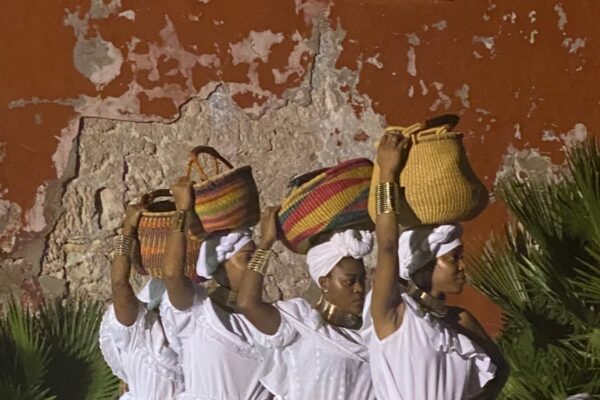 Four women dressed in long white ruffled dresses carry baskets on their heads.
