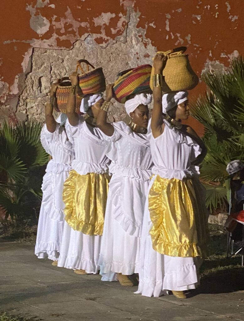 Four women dressed in long white ruffled dresses carry baskets on their heads.