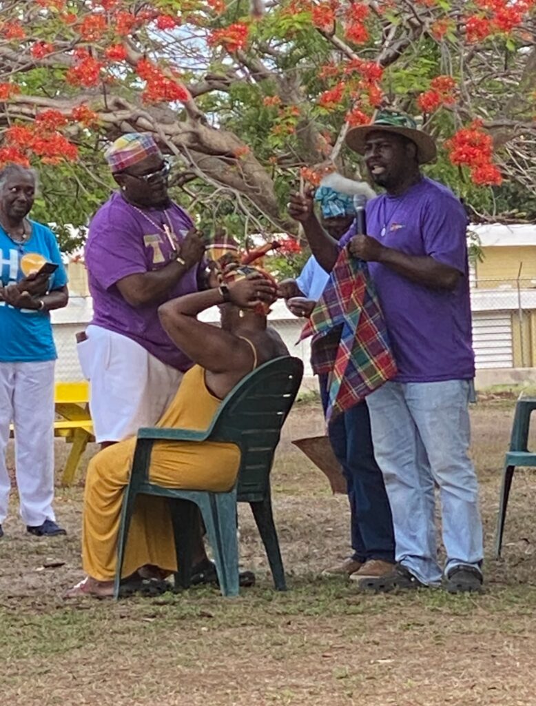 A woman sits outside under a flowering tree. Two men demonstrate how to tie a madras headscarf on her head.
