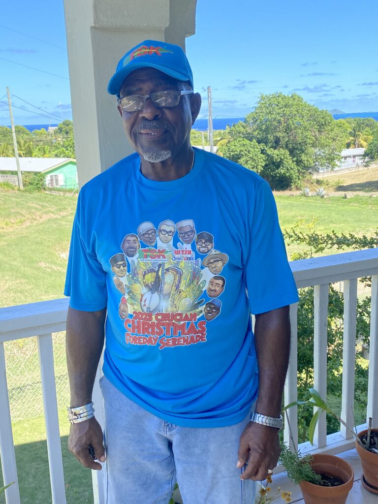 A man stands on a covered porch balcony with the blue sea visible in the background.