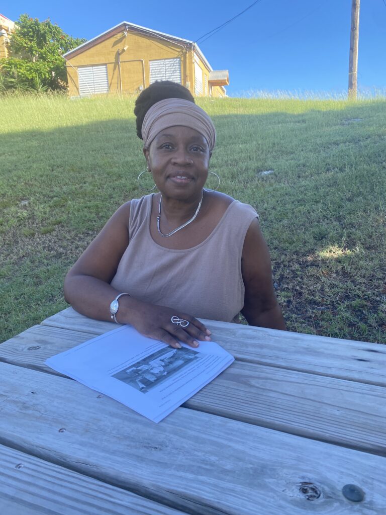 A woman sits at a picnic table with a sloping lawn of grass and bright blue sky behind her.