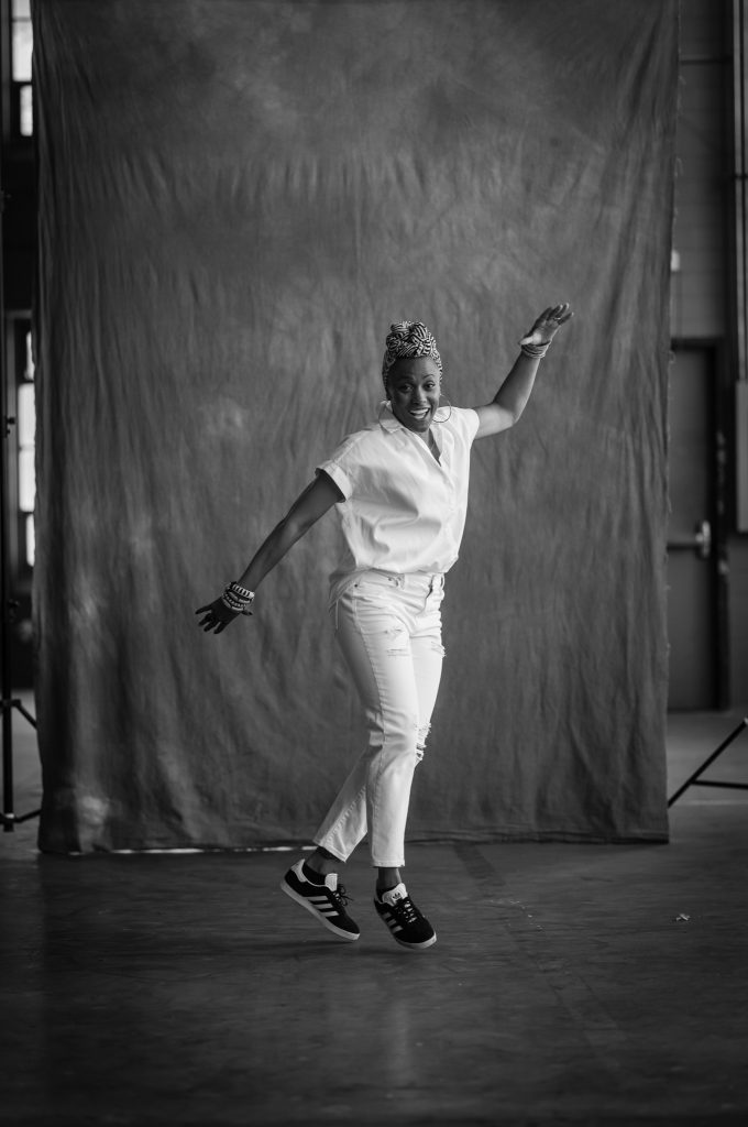 Black and white image of Michele Byrd-McPhee dress in white dancing in front of a studio backdrop.