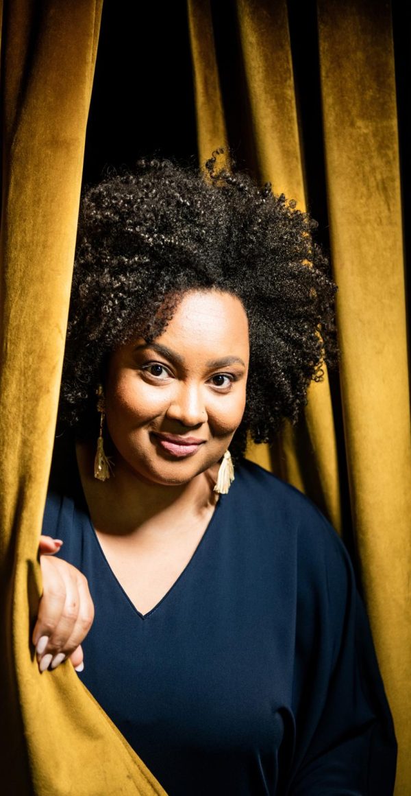 Lucibela looks around the edge of a gold stage curtain.