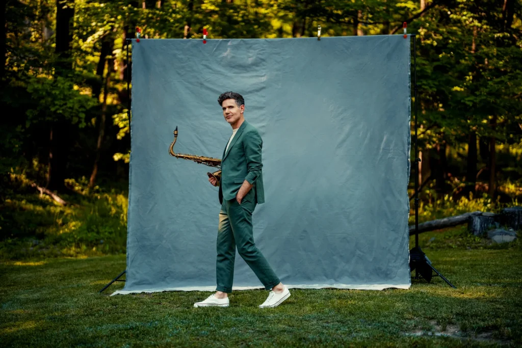 Josh holds a sax and poses outside in front of a blue background.