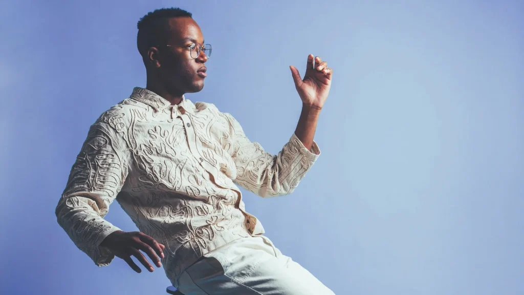 A man sits on a stool and poses in front of a blue background.